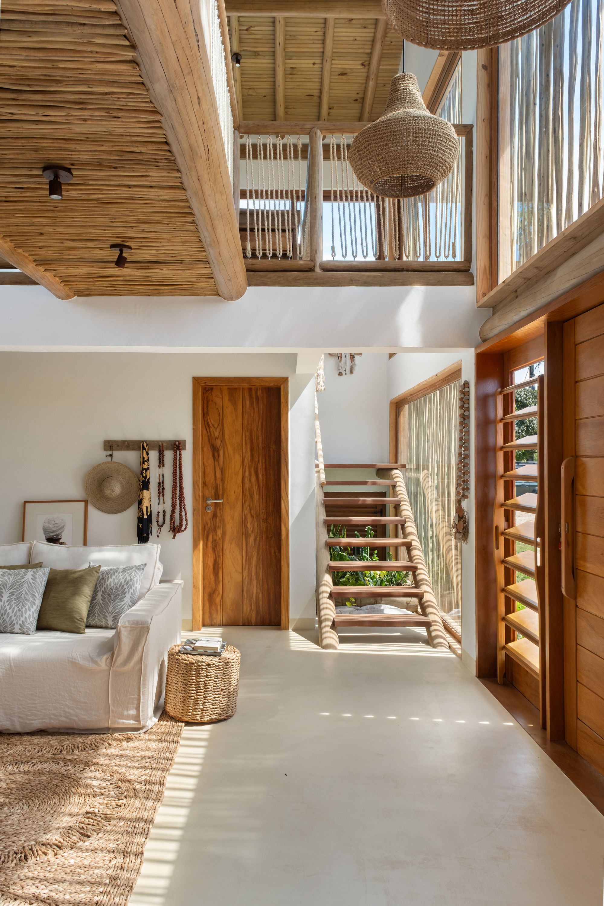 Double-height entry: the rope balustrade of the footbridge above, a woven pendant, and a timber stair in striped light.