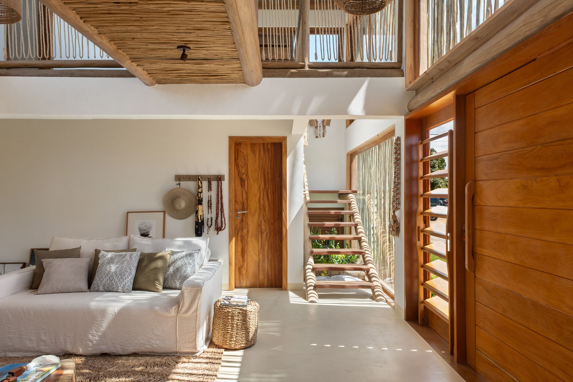 Living room toward the stair: linen sofa, a straw hat and beads on the wall, a wooden door, and the stair lit through the stick screen.