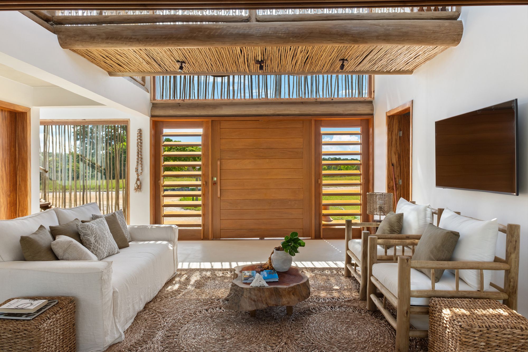 Living room facing the front door: linen sofa, timber armchairs, a tree-trunk coffee table on a jute rug, and the pivot door between louvred panels.