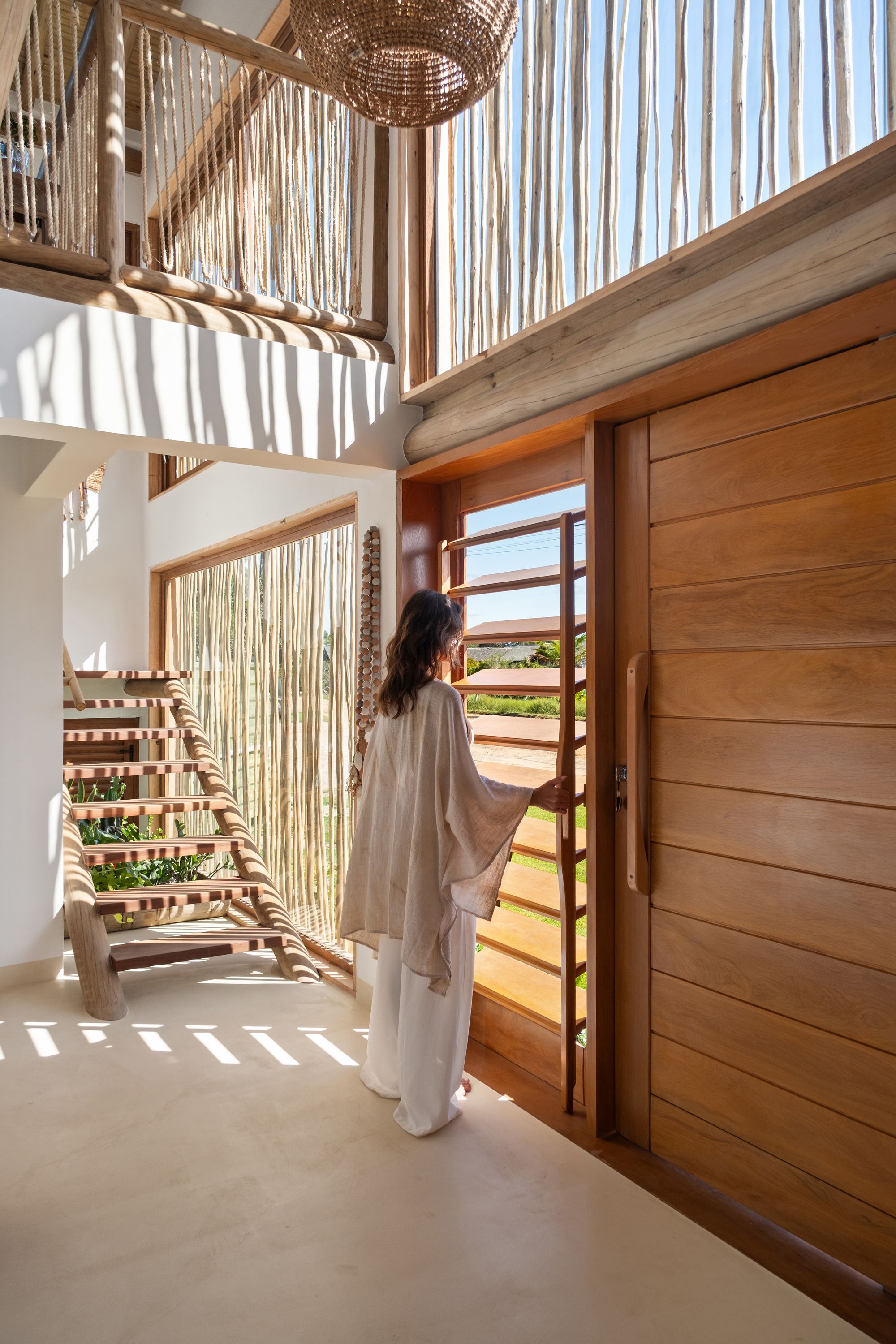 A woman opens a louvred panel beside the pivot door; striped shadows fall across the floor and the timber stair.