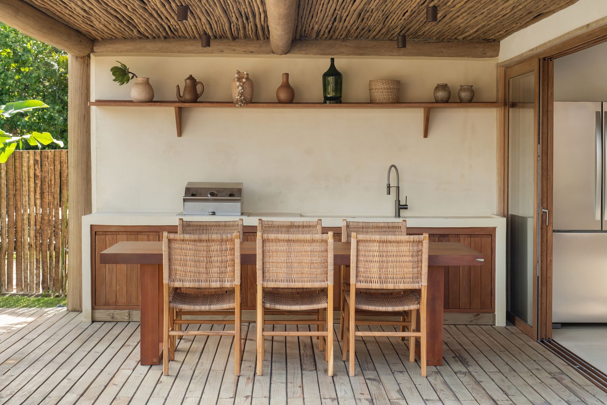 Outdoor kitchen: a cream counter with grill and sink, a shelf of clay vessels, and a timber table with woven chairs.