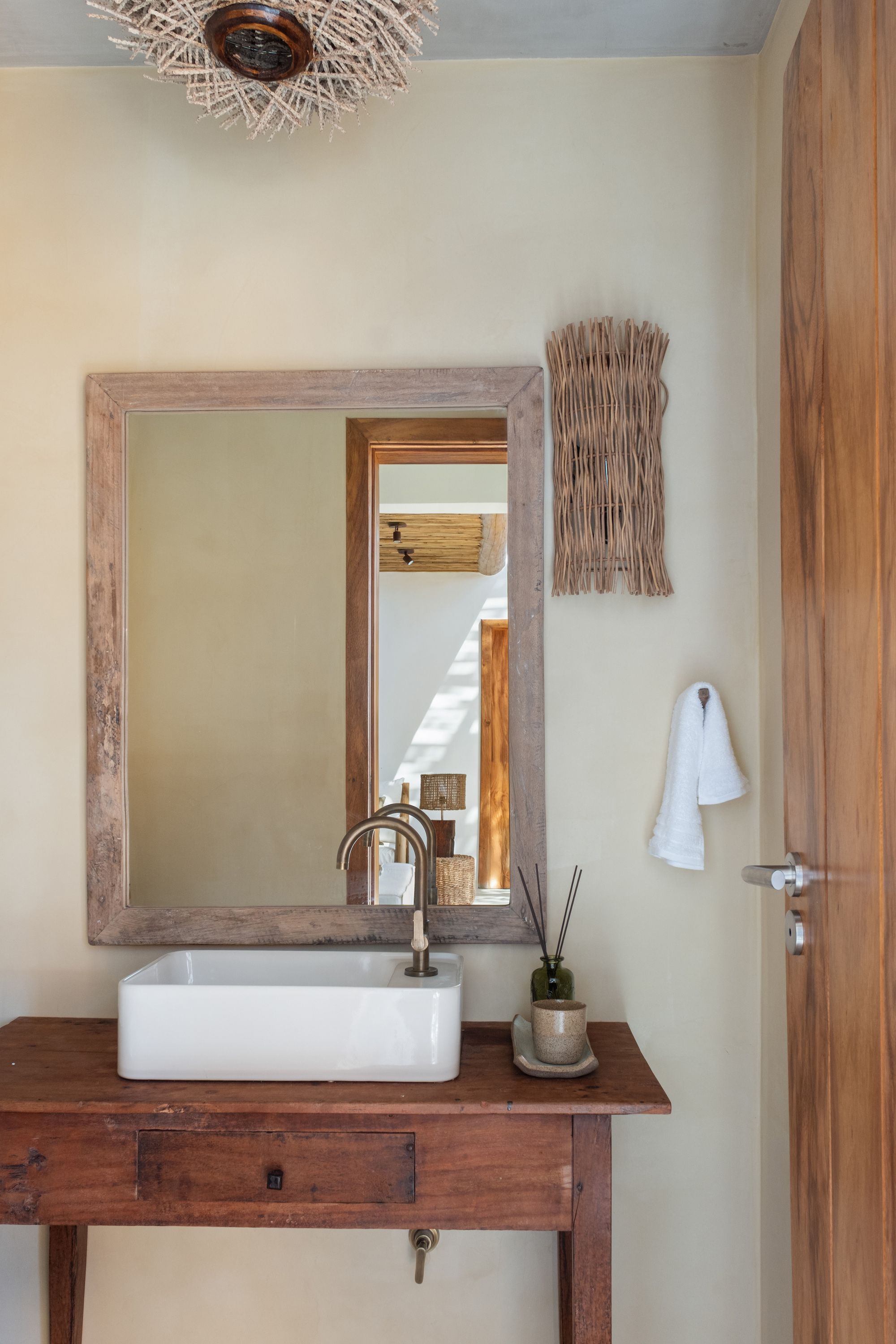 Guest washroom: a white basin on an old timber table, a weathered wood mirror, and a woven wall lamp.