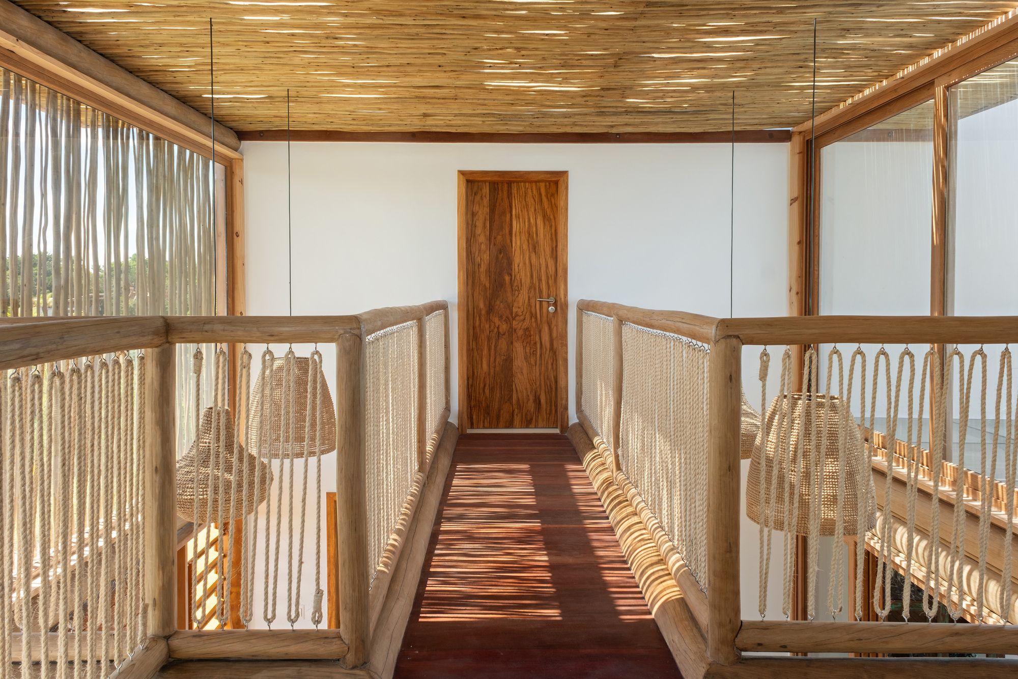 The footbridge: a dark timber walkway with round-log rails and looped sisal rope balustrades, under a stick ceiling, leading to a suite door.