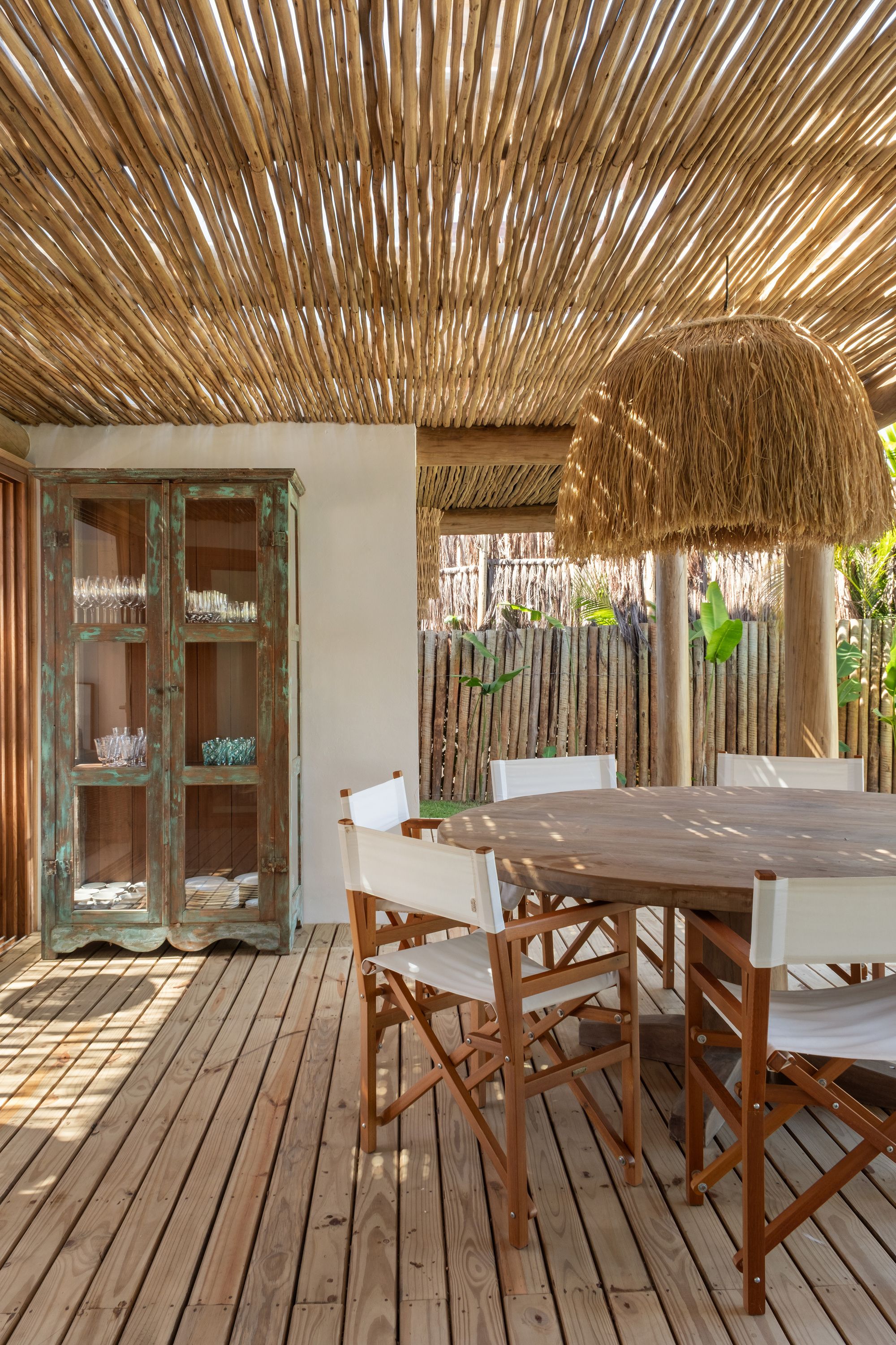 Pergola dining area: an antique turquoise cabinet, a raffia pendant, and striped light falling on the deck; the timber-stake fence beyond.