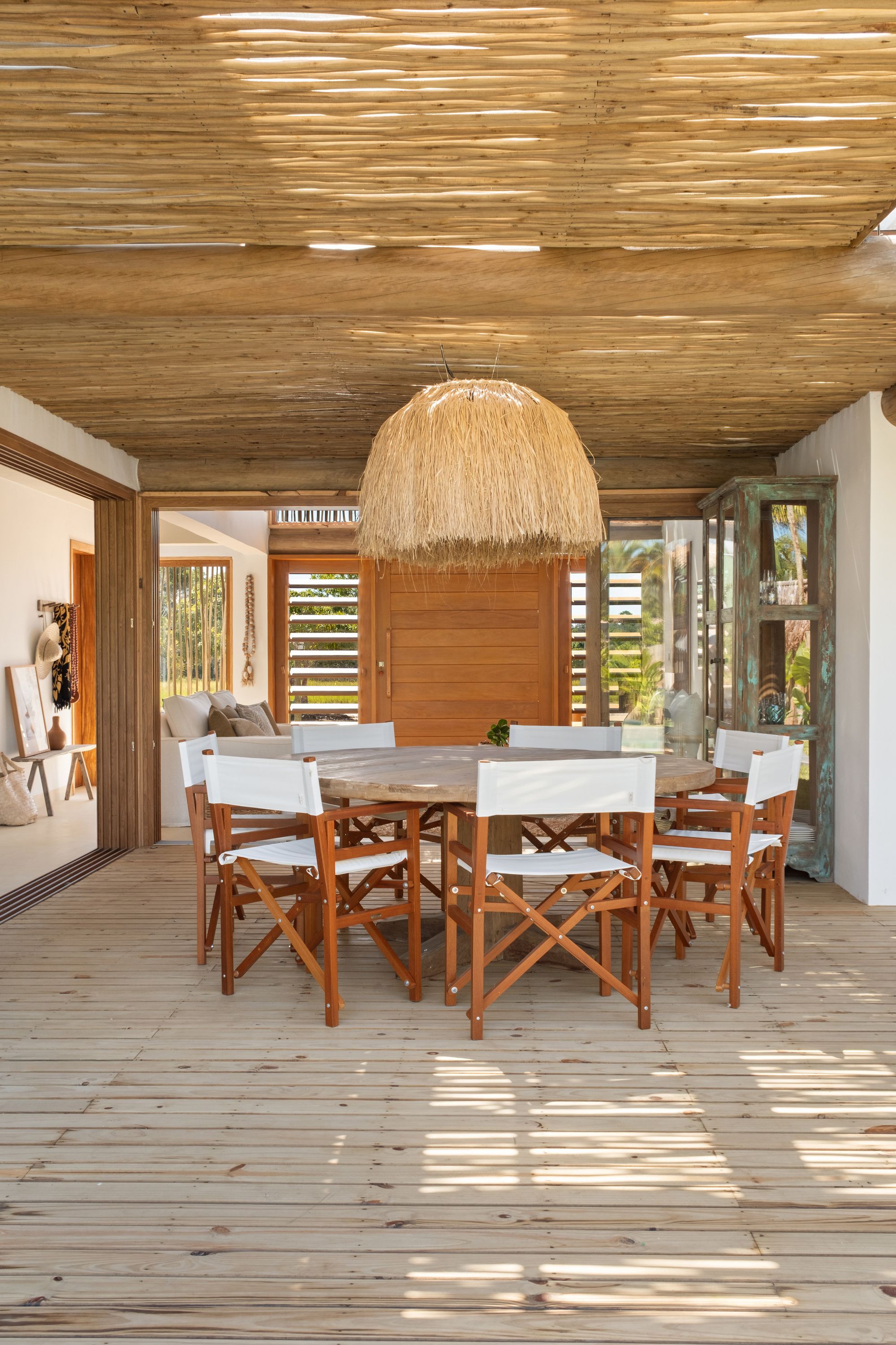 Round dining table with white director’s chairs under the stick-covered pergola; a large raffia pendant hangs above and the pivot door is visible beyond.