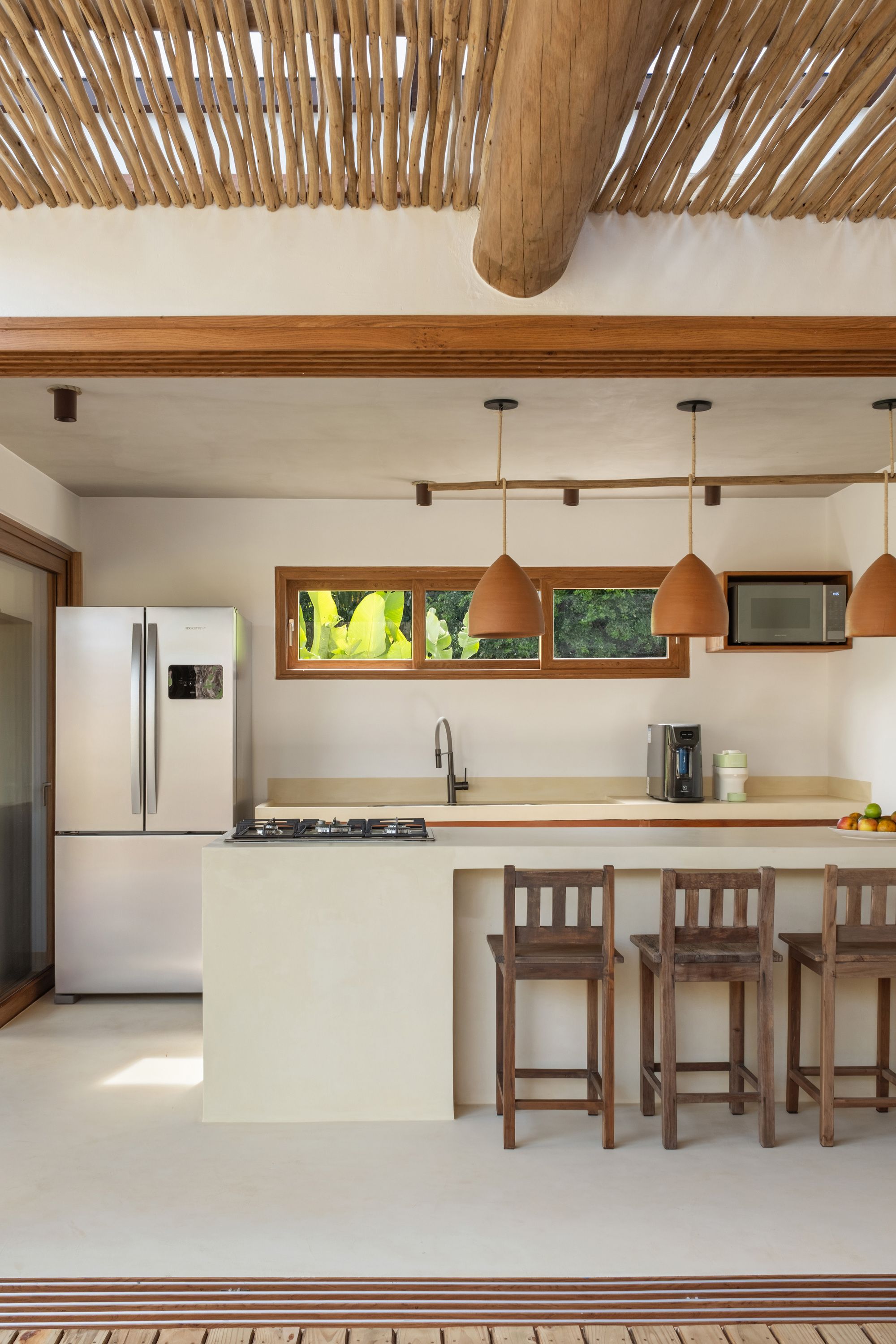Kitchen island in cream microcement with wooden stools, terracotta pendants on a branch rail, and a round log beam overhead.