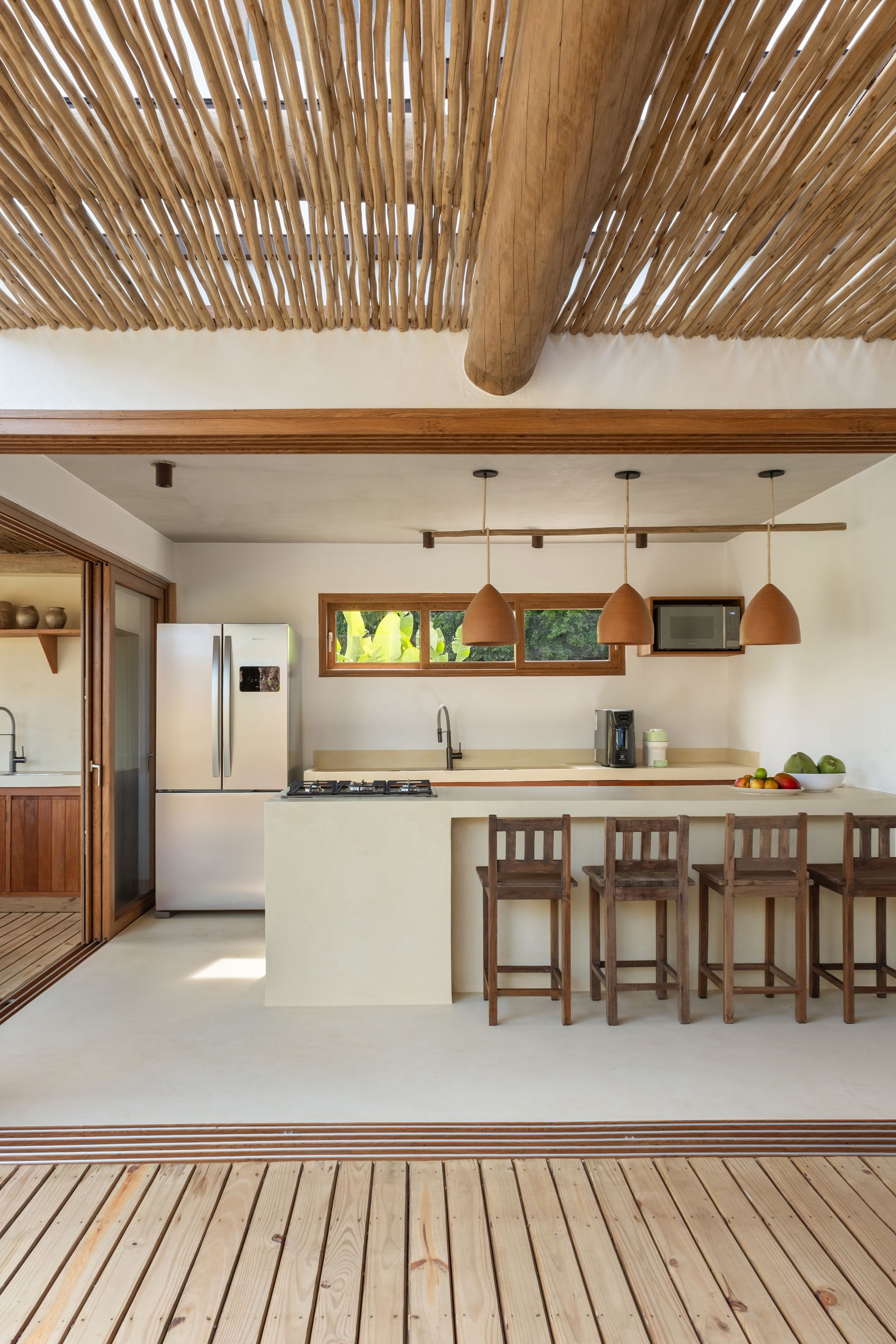 Kitchen opened fully to the deck: a cream microcement island, wooden stools, terracotta pendants, and the pergola’s stick ceiling above.