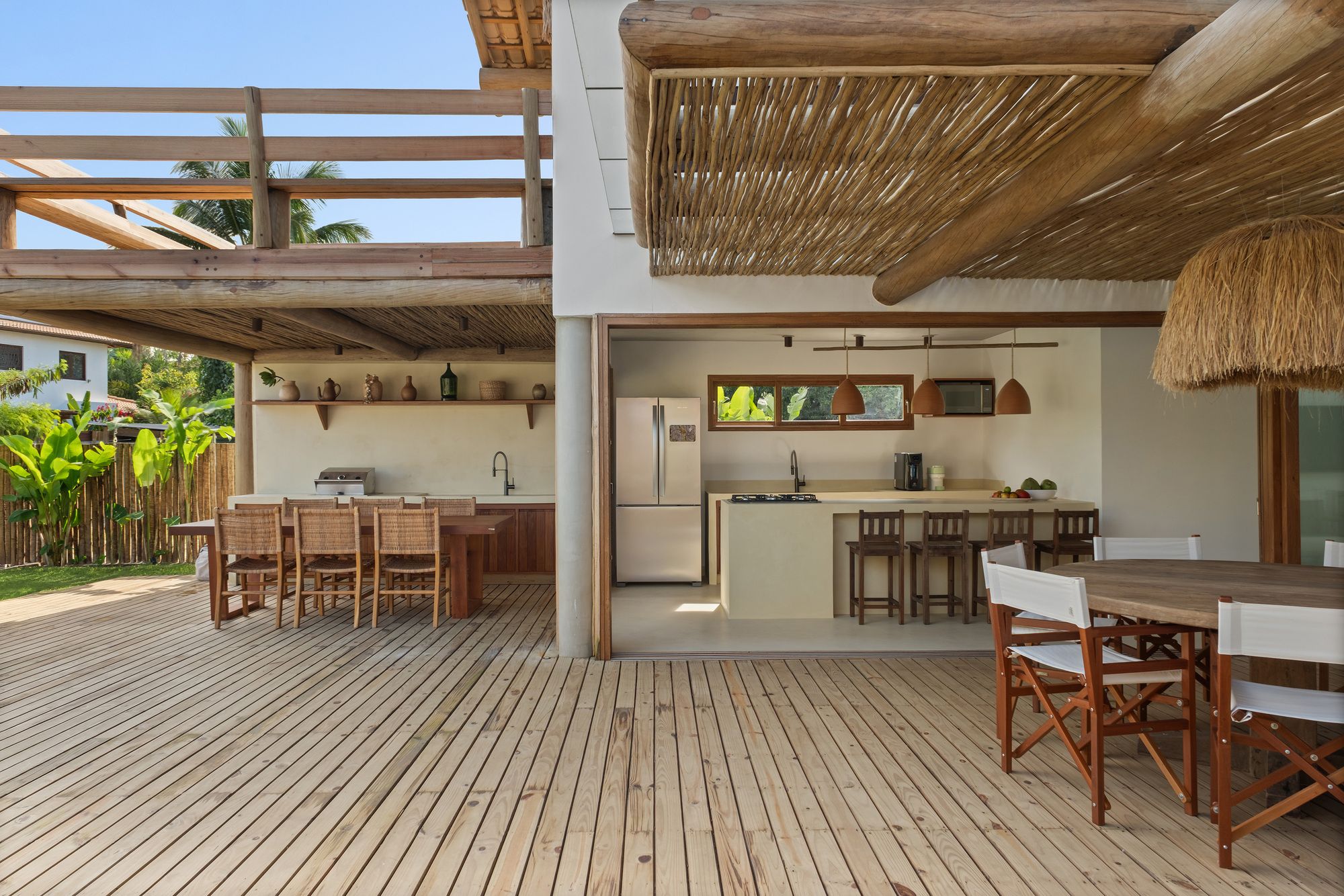 The outdoor kitchen with a long timber table, the kitchen behind an open glass corner, and the dining table under the pergola.