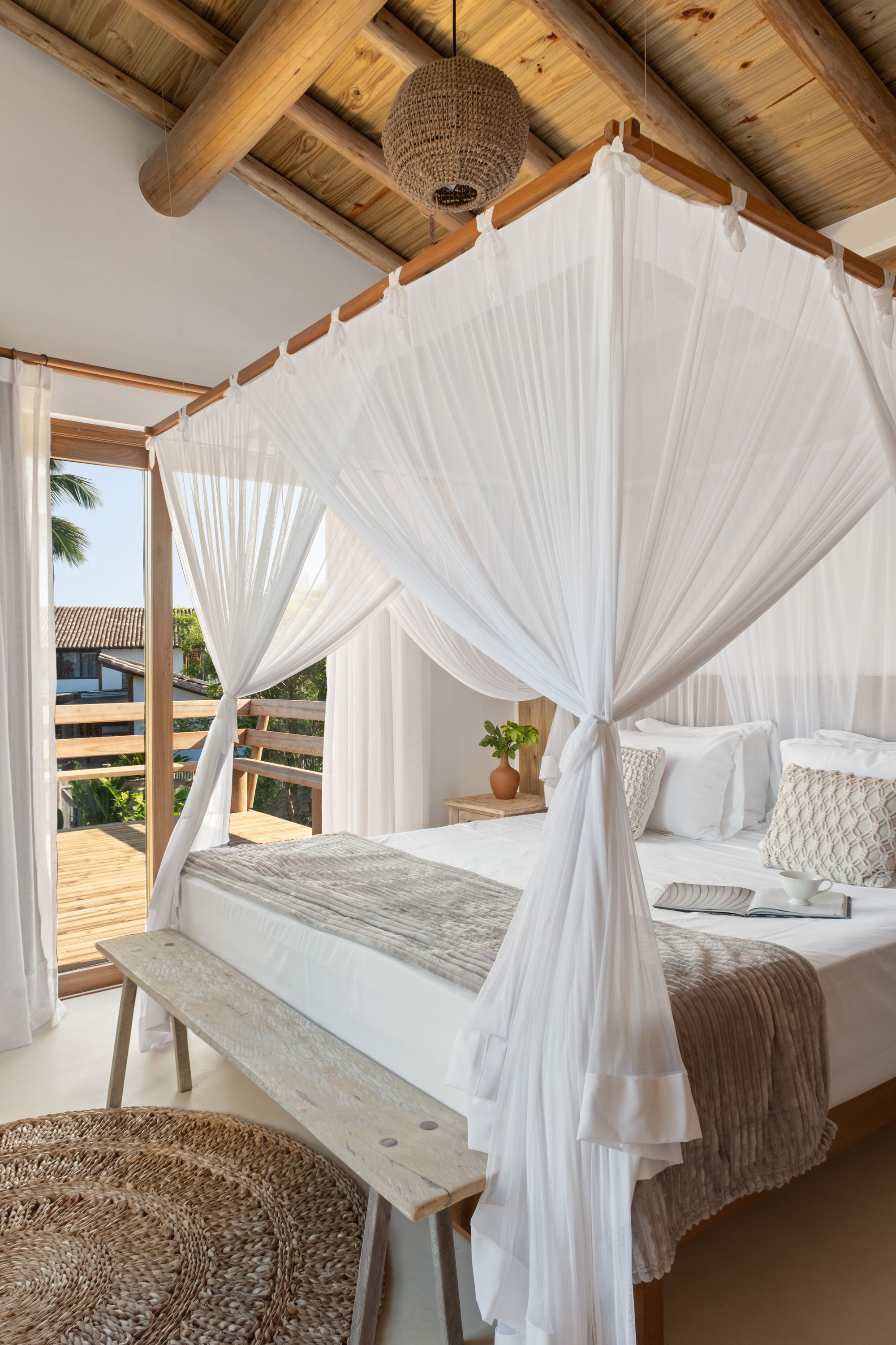 Upper suite: a bed under a white mosquito net, round-log rafters, and a door to a timber-railed balcony.
